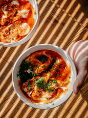 Hand gripping wooden bowl filled with traditional turkish shakshuka, garnished with yogurt, tomato sauce, spices, dill, beside companion bowl under soft natural illumination. Bowl of turkish shakshuka