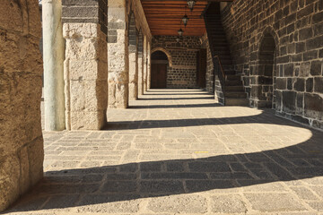 Architectural perspective of traditional stone corridor with arched columns and wooden ceiling beams, casting long shadows across sunlit stone floor