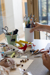 A well-lit craft table with an assortment of art and crafting supplies, including brushes, measuring tape, and jewelry tools, suggesting a creative project in progress. A hand reaches for a brush.