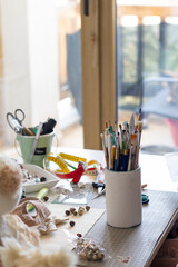 Window-lit craft desk, overflowing with brushes, beads, scissors, and natural elements. A scene of vibrant creativity.
