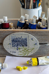A close-up of an artist's workspace featuring tubes of paint, brushes in a holder, and a decorative plaque with a lavender field scene. Yellow paint is squeezed onto paper, ready for use.