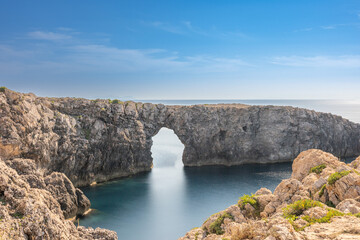 Pont d'en Gil in Menorca viewed from the front, with silky water from long exposure, rocky foreground, and a blue sky in the background.