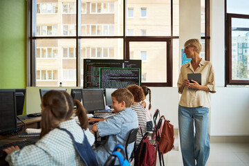 Caucasian middle aged woman holding tablet standing near group of diverse children sitting at desks using desktop computers in classroom setting, teacher observing students working