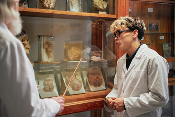 Young adult Caucasian man wearing lab coat listening attentively to senior man explaining anatomical specimens in glass jars inside scientific museum or laboratory setting