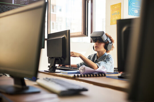 Teenager boy with curly hair using virtual reality headset and headphones while interacting with computer monitor in classroom setting, engaging with digital learning technology