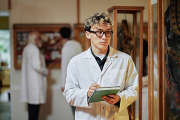 Young adult Caucasian man wearing glasses standing in laboratory holding notepad and writing, with two middle aged men in lab coats discussing in background, scientific setting