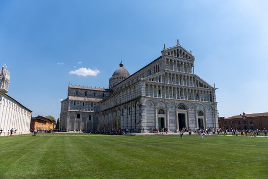 Pisa Cathedral in Italy, showcasing Romanesque architecture with ornate fa&ccedil;ade, arcades, and a central dome. Surrounded by green lawns and visitors in Piazza dei Miracoli, a UNESCO World Heritage Site