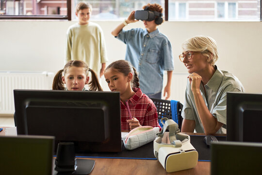 Caucasian senior woman supervising two preteen girls working at computer while two boys in background using virtual reality headset in modern classroom technology lesson - Powered by Adobe