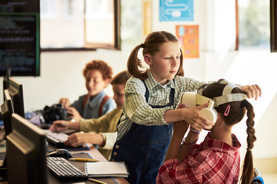 Caucasian girl helping another Caucasian girl put on virtual reality headset in classroom while two multiethnic boys using computers in background, children engaging with technology together