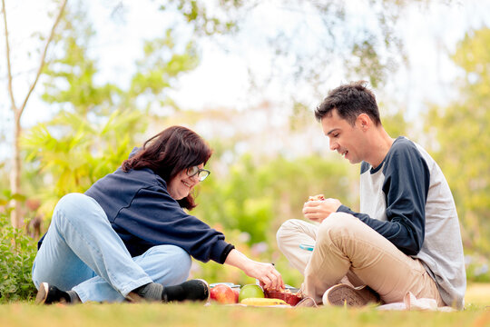 Mother and adult son enjoying an outdoor breakfast on the grass, sharing fruit, pie, and coffee in a relaxed and happy moment.