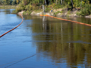Orange oil containment booms on a river, used to prevent the spread of oil pollution or to collect floating debris. A riverbank and trees are visible in the background. © Andrey