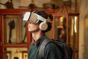 Young adult Caucasian man wearing virtual reality headset and headphones standing in museum environment with backpack, exploring digital content through immersive technology