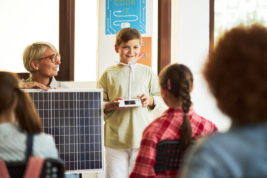 Caucasian middle aged woman holding solar panel standing next to Caucasian boy presenting small device to group of teenagers, sitting and listening during classroom lesson - Powered by Adobe