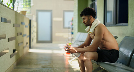 Indian gen z young adult male man guy sitting on chair in hallway hold bottle. Asian relax boy take break after sports workout spend gym time look sad upset outside corridor area