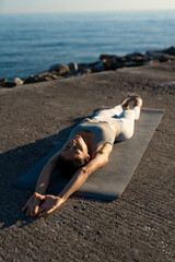 Relaxed woman lying on a yoga mat, stretching her arms above her head, enjoying the ocean view under a soft evening light