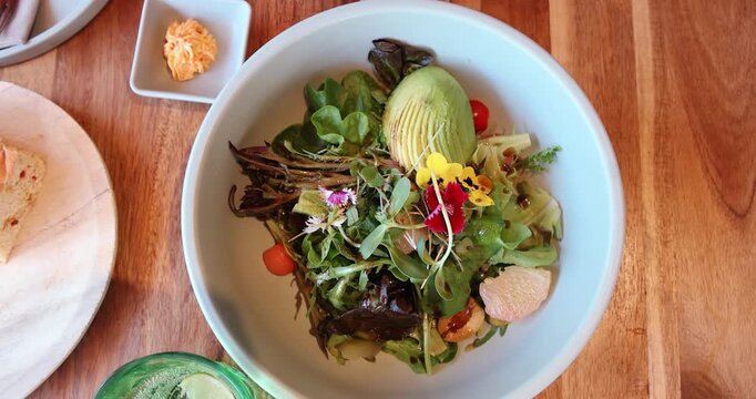 Salad bowl with avocado, mixed greens, edible flowers, cherry tomatoes on wooden table. Healthy meal, fresh produce, organic eating concept.
