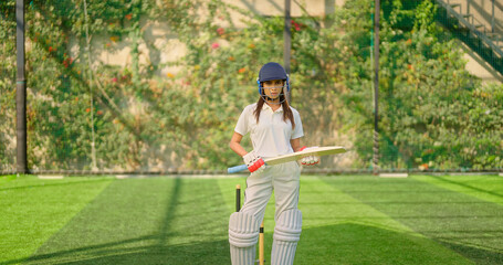 Indian young adult gen z sports woman lady standing at outdoor ground day wear white uniform and helmet look camera. Asian female girl player hold bat staring cam play do casual pose