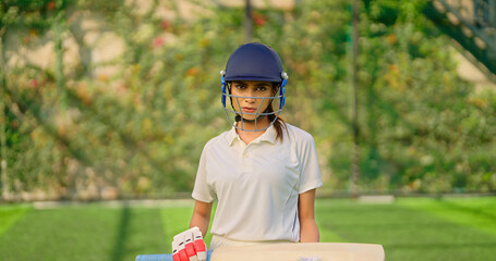 Indian gen z athlete sports woman lady standing staring cam play ground wear white uniform and protective helmet look camera Asian cricket player female girl hold bat day time at match play ground
