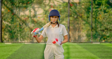 Indian gen z athlete sports woman lady standing at outdoor ground day wear white uniform and helmet look camera Asian one cricket player female girl hold bat ball staring cam do casual pose 