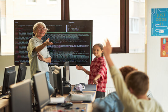 Senior Caucasian woman teaching coding to group of children in computer classroom, girl standing near interactive screen displaying programming code, students raising hands - Powered by Adobe