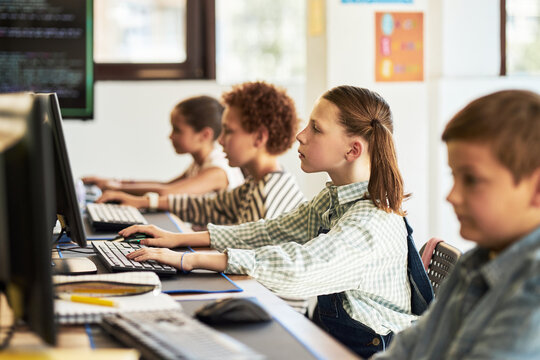 Group of diverse children sitting at desks using desktop computers in classroom, focused on typing and looking at monitors, learning technology skills during school lesson