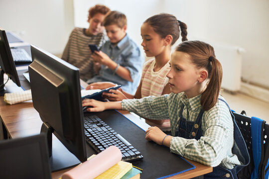 Group of Caucasian and multiethnic children sitting at desk using desktop computers and smartphone in classroom, focused expressions showing teamwork and learning technology skills together