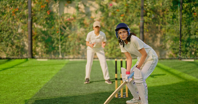 Indian gen z young adult sports woman girl standing at outdoor match play ground wear gloves white uniform and protective helmet Asian athlete cricket player female lady hold bat do practice day time