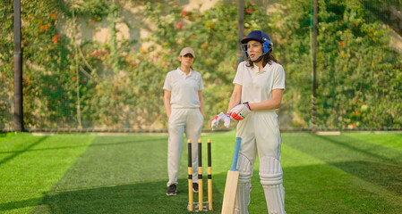 Fototapeta premium Indian gen z young adult sports girl at match play ground wear gloves white uniform leg guard and helmet Asian two cricket player female lady standing hold bat do casual pose ready for match day