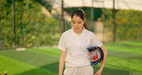 Indian gen z relax sports teen age girl woman standing outdoor play ground wear white uniform hold protective helmet and hand gloves Asian cricket player female take break rest time at match ground