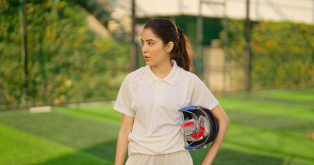 Indian gen z relax sports teen age girl woman standing outdoor play ground wear white uniform hold protective helmet and hand gloves Asian cricket player female take break rest time at match ground