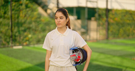Indian gen z relax sports teen age girl woman standing outdoor play ground wear white uniform hold protective helmet and hand gloves Asian cricket player female take break rest time at match ground