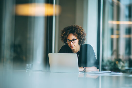 Focused woman working on laptop in modern office, wearing glasses with curly hair, showcasing concentration and professionalism.