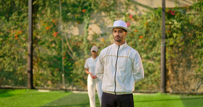 Indian young adult gen z bowler woman standing outdoor match play ground wear uniform cap Asian female lady throwing ball do practice umpire man standing watch player cricket game - Powered by Adobe