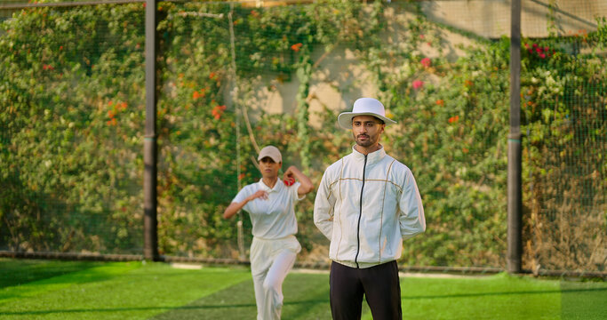Indian young adult gen z bowler woman standing outdoor match play ground wear uniform cap Asian female lady throwing ball do practice umpire man standing watch player cricket game