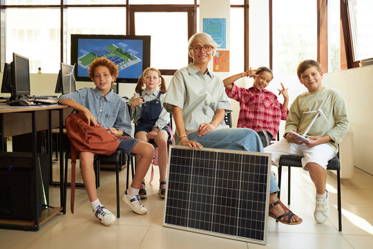 Caucasian middle aged woman sitting with diverse group of children in classroom, holding solar panel, children smiling and gesturing, computer monitors visible in background