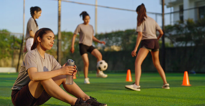 Indian happy athlete woman lady player group play football game kicking soccer ball enjoy team young adult gen z tired girl female sitting take break practice health care hold bottle drink water