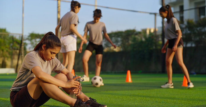 Indian athlete woman lady player group play football game kicking soccer ball enjoy team young adult gen z tired girl female sitting take break practice health care hold bottle drink water - Powered by Adobe
