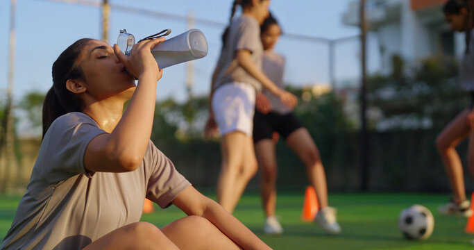 Indian happy athlete woman lady player group play football game kicking soccer ball enjoy team young adult gen z tired girl female sitting take break practice health care hold bottle drink water