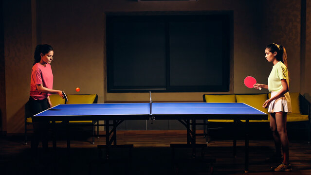 Indian happy young adult gen z two athlete player woman hold racket play table tennis passing ball indoor dark room. Asian sporty girl lady do sports training enjoy ping pong game have fun joy	