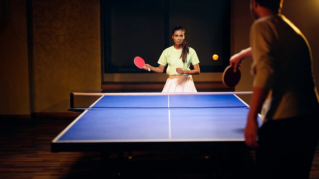 Indian happy young adult gen z athlete player woman hold racket play table tennis male man passing ball indoor dark room Asian sporty girl lady do sports training enjoy ping pong game have fun joy	 - Powered by Adobe