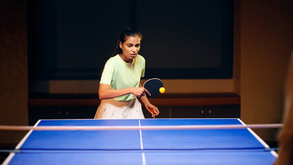 Indian young adult gen z athlete player woman hold racket play table tennis passing ball indoor dark room Asian sporty girl lady do sports training enjoy ping pong game have fun joy	