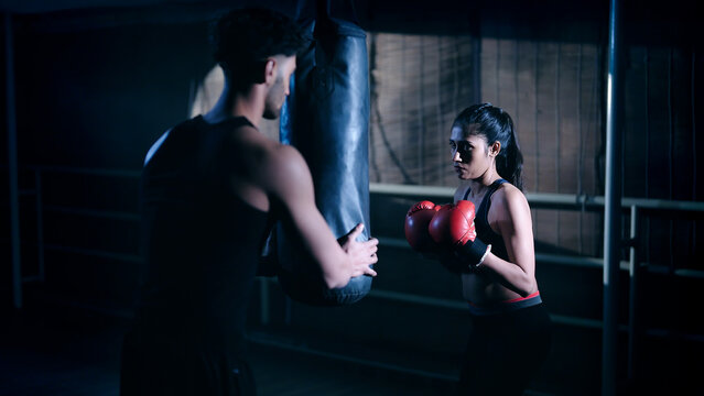 Indian young adult gen z angry fit boxer woman lady wear gloves do hand fist boxing punch beat on bag indoor dark room place modern trainer guy sports female girl learn self defense martial art skill - Powered by Adobe