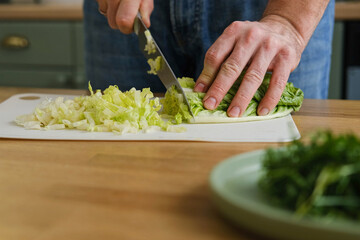 Handsome bearded man is preparing green fresh vegan salad in the kitchen at home. Male cutting vegetables, lettuce with knife on wooden chopping board, cooking vegetarian meal. Tasty healthy diet food