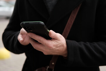 Handsome man wearing black coat and brown leather bag walking down on the city with mobile phone in hand. Stylish bearded middle-aged male using smartphone at urban street. Businessman going on job.