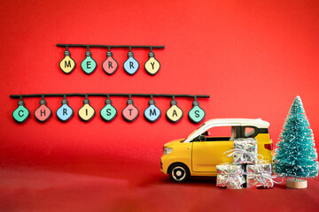 Yellow car with Christmas presents and tree on red background. Merry Christmas lettering in light bulbs in the background