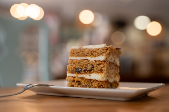 Close-up of a delicious slice of homemade carrot cake with cream cheese frosting, served on a white plate with a fork on a wooden table, captured in a cozy cafe with warm lighting