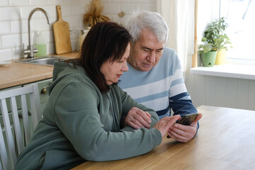 Old couple using a phone together while relaxing in the kitchen at home. Elderly man and woman spending leisure time browsing online with a smartphone. Happy senior married couple typing, learning