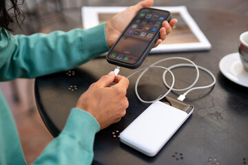 Close-up of a person connecting a smartphone to a white power bank using a charging cable at a café table, showing modern mobile technology and lifestyle on the go
