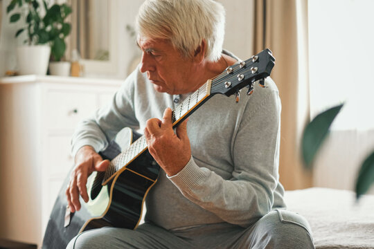 Senior man is playing guitar. Elderly male sitting at sofa and play music. Portrait of a gray haired mature 60s person with acoustic guitar, he is learning to play. Enjoying retirement life at home. - Powered by Adobe