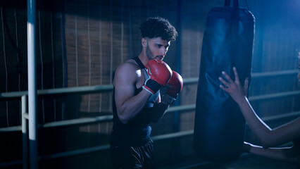 Indian young adult gen z angry fit boxer man guy wear gloves do hand fist boxing punch beat hit on bag indoor dark class room place modern sports smart male boy learn self defense martial art skill